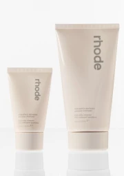 Two pale beige rhode skincare squeeze tubes in mini and full sizes standing on a reflective white surface against a white background.