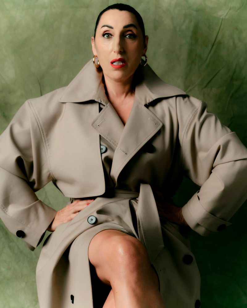 Rossy de Palma wearing an oversized beige trench coat with hands on hips against an olive green textured backdrop.