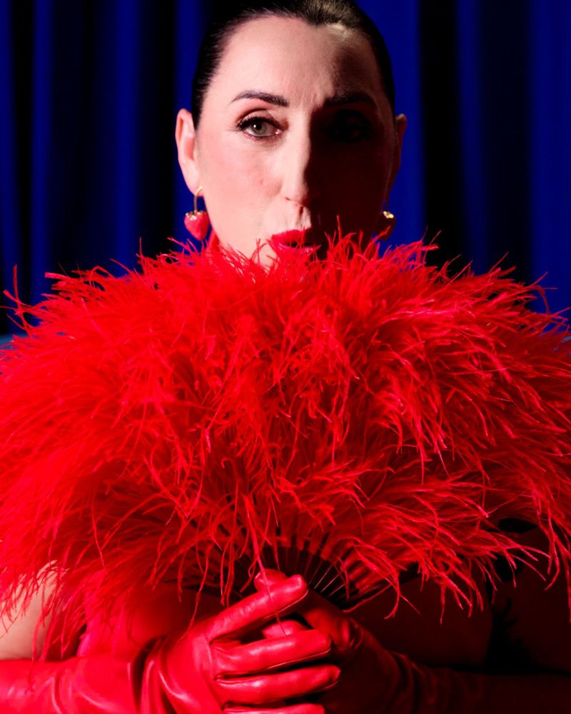 Rossy de Palma holding a voluminous red feather fan while wearing red leather gloves against a rich blue curtain backdrop.