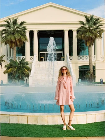 Model wearing a pink ruffle mini dress and platform espadrille wedges standing before a large fountain and neoclassical building.