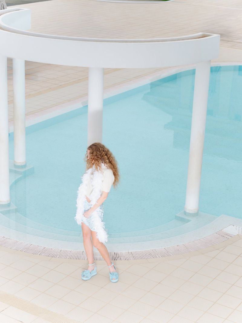 A model with long wavy hair wears a white feathered stole and light blue platform shoes beside a curved architectural pool.