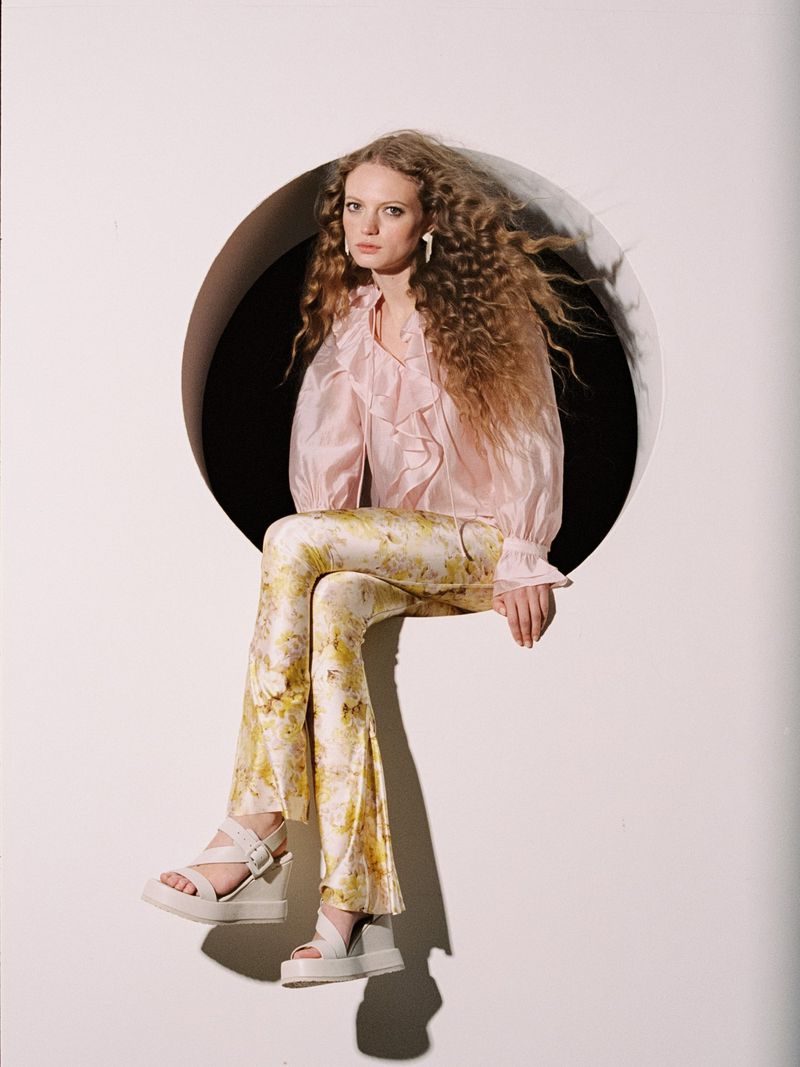 Model with curly hair seated in a circular wall cutout wearing a ruffled pink blouse, floral trousers, and white platform sandals.