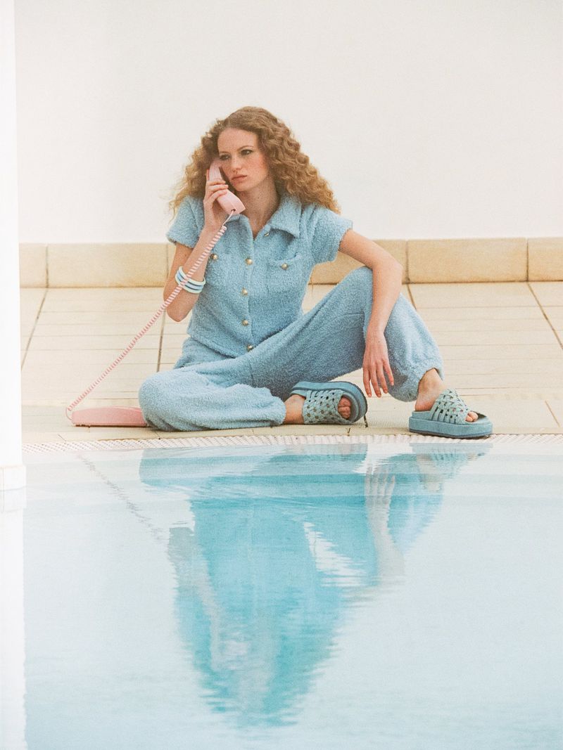 Woman in a light blue textured jumpsuit and platform slides sits by a pool holding a pink telephone receiver, reflected in the water.
