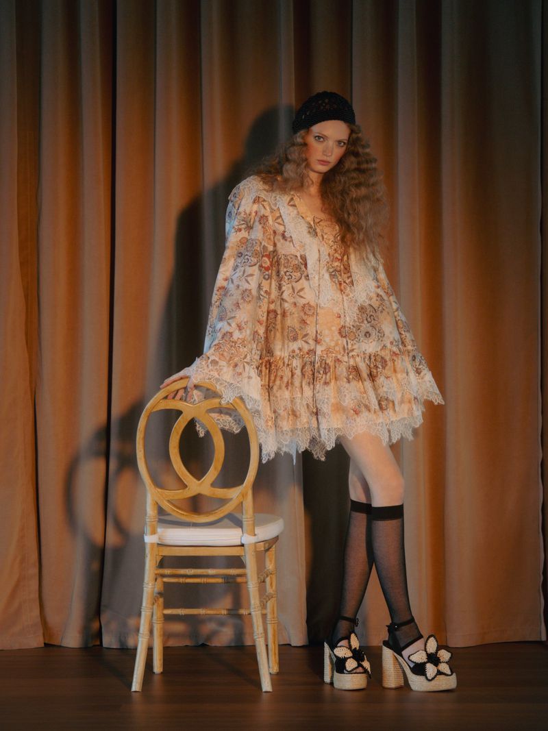Model in a floral lace mini dress and statement platform sandals leaning on an ornate wooden chair before draped curtain backdrop.