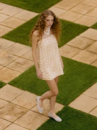 Woman in a pale mini dress and white woven flats standing on a checkerboard lawn of grass and stone pavers.