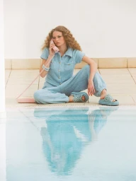Woman in a light blue textured jumpsuit and platform slides sits by a pool holding a pink telephone receiver, reflected in the water.