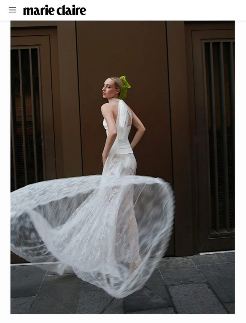 Blonde woman in a white halterneck dress spinning with a flared lace skirt in front of a bronze building facade.