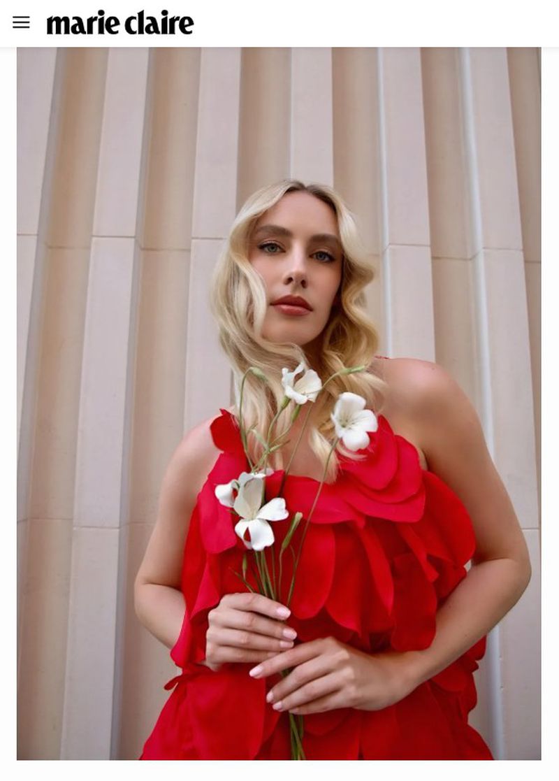 Blonde woman in a ruffled red dress holding white flowers against a vertically ribbed architectural wall.