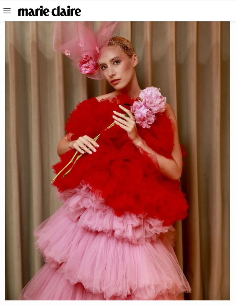Fashion model in a voluminous red and pink tiered tulle gown holding pink peonies against pleated neutral fabric curtains.