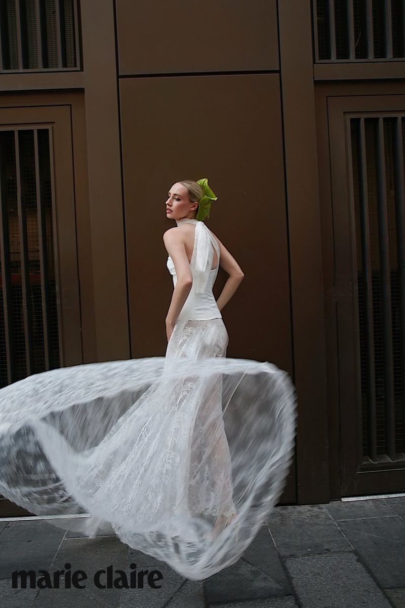 Model spinning in a sheer white lace dress with a green hair bow in front of a dark building exterior.