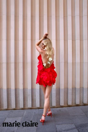 Blonde woman in a red backless petal dress with flowers along her spine and matching red heels against a fluted beige wall.
