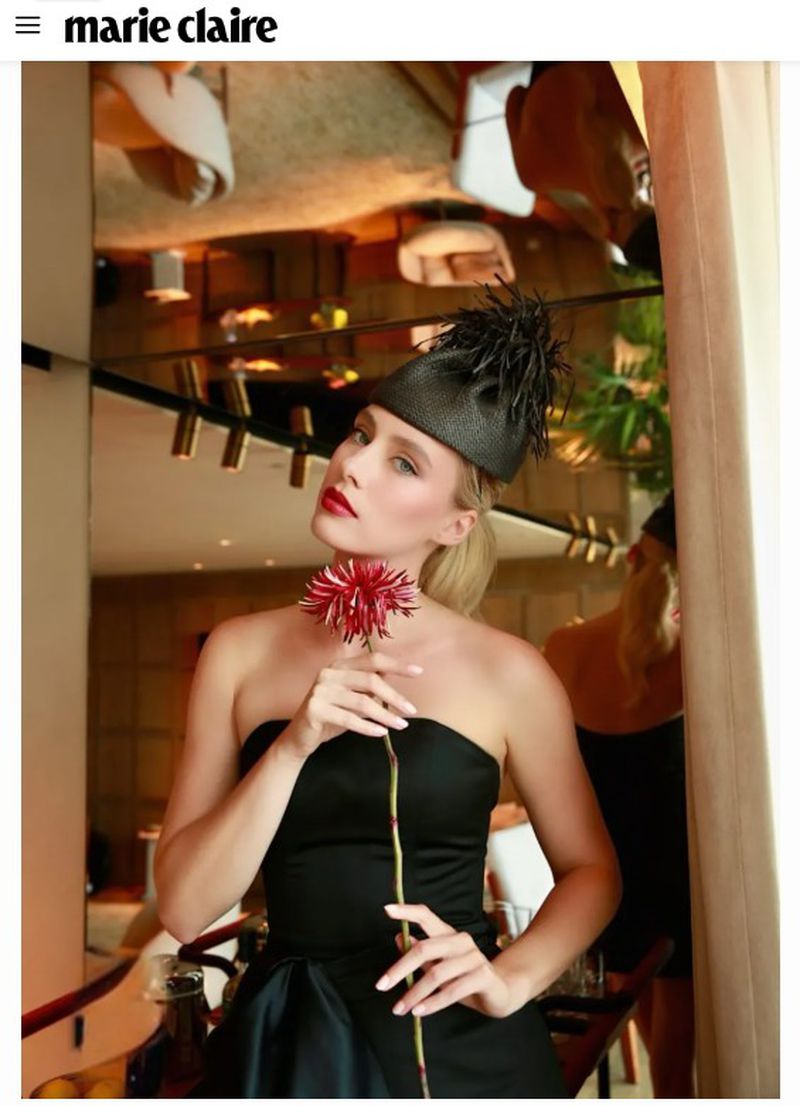 A blonde woman in a strapless black dress and feathered pillbox hat poses holding a long-stemmed red flower in front of a mirror.