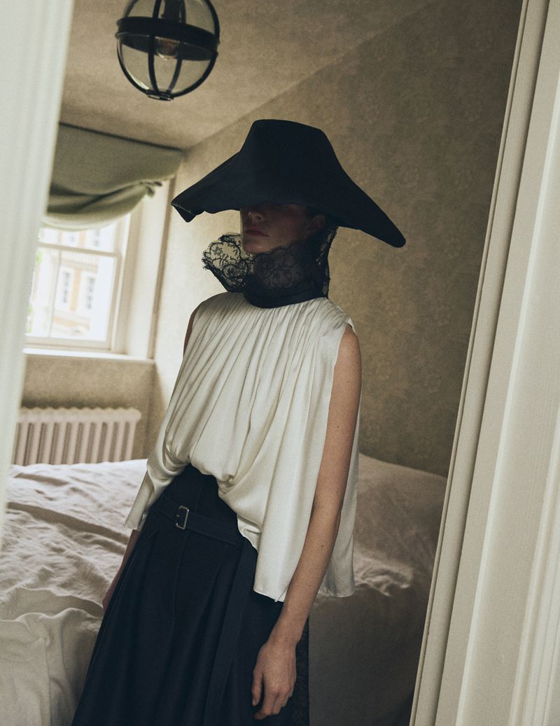 Model Carolina Burgin wearing an oversized black sculptural hat, lace collar, and draped ivory top in a bedroom for Vogue Scandinavia.