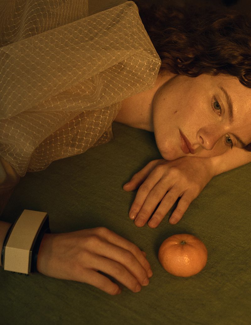 Model Carolina Burgin resting her head near an orange on a green surface, wearing a sheer netting top and wide cuff bracelet.