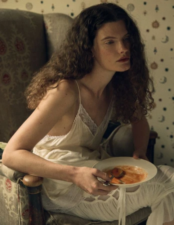 Model with long curly hair wearing a white slip dress seated in an armchair holding a bowl of soup for Vogue Scandinavia Victorian Era