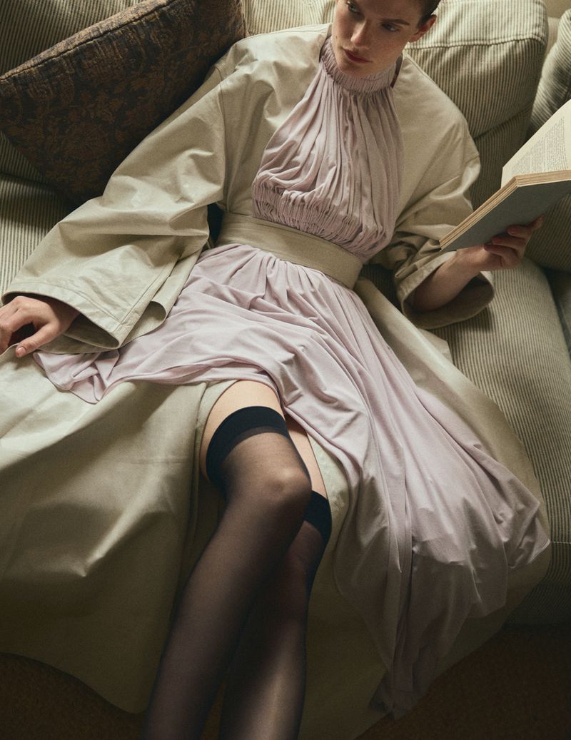 Model in a vintage-style high-neck pleated dress and beige coat holding a book while seated on a striped sofa.