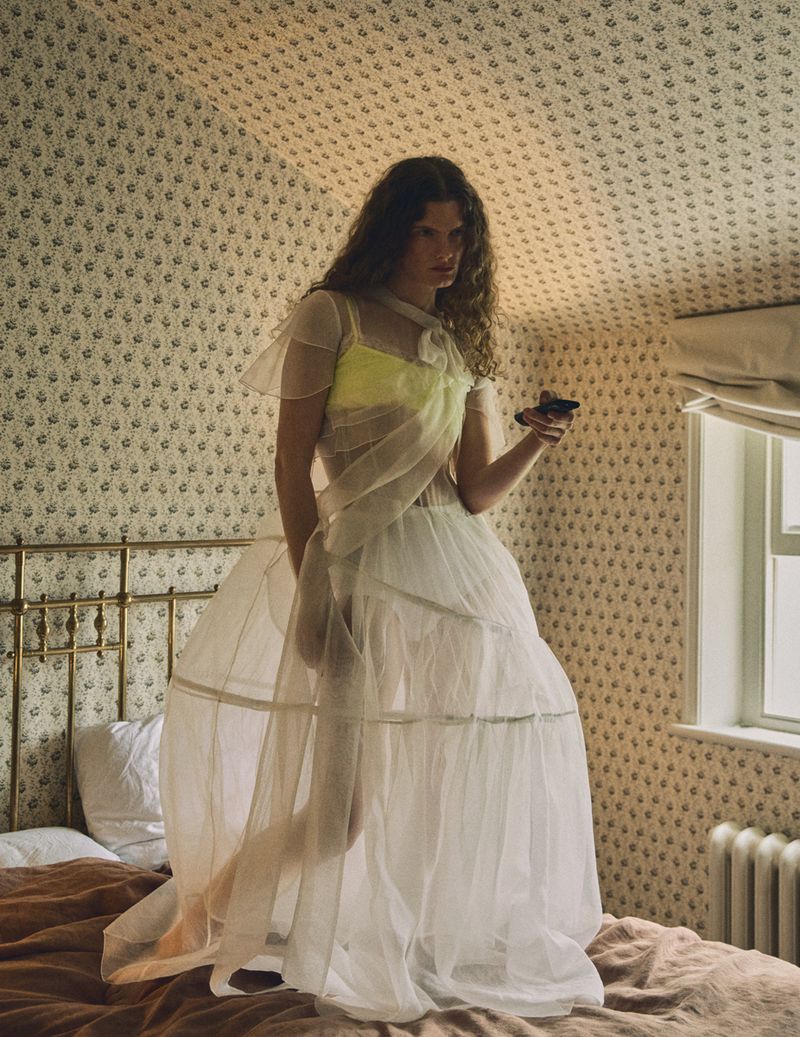 Model standing on a bed in a sheer white crinoline gown holding a smartphone in a vintage bedroom with floral wallpaper.