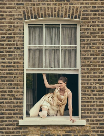 Model Carolina Burgin crouching inside a white sash window of a brown brick building in a draped cream dress.