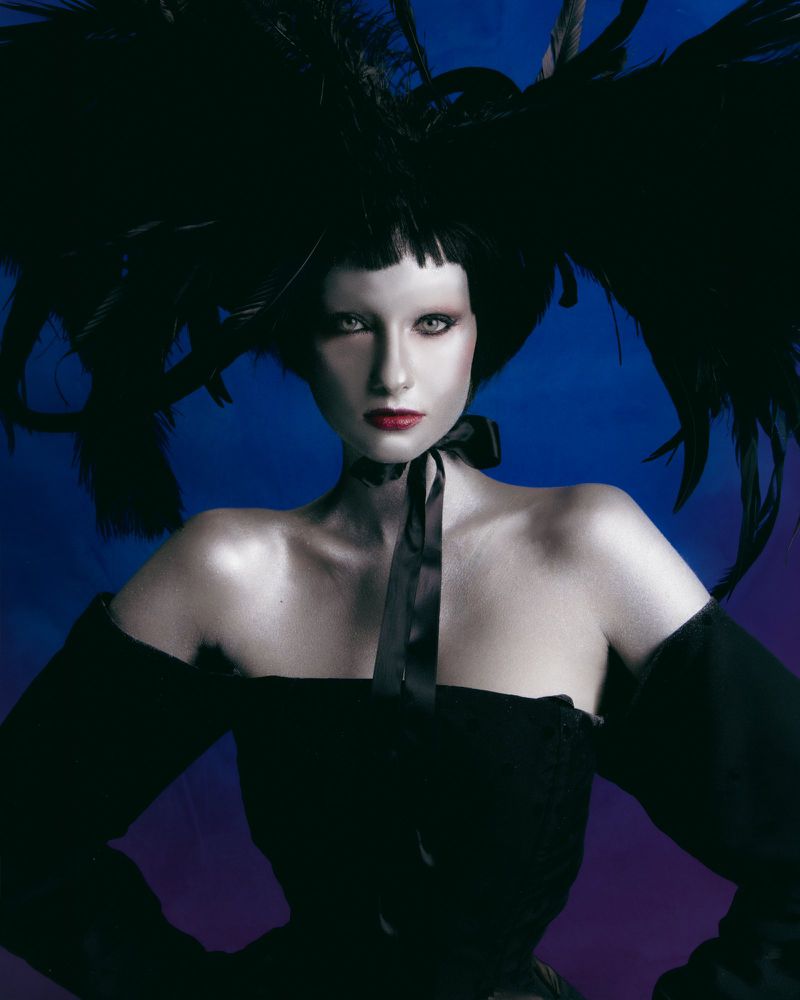 Model wearing a dramatic black feathered headpiece and an off-the-shoulder black dress against a blue gradient backdrop.