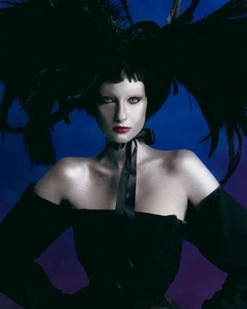 Model wearing a dramatic black feathered headpiece and an off-the-shoulder black dress against a blue gradient backdrop.