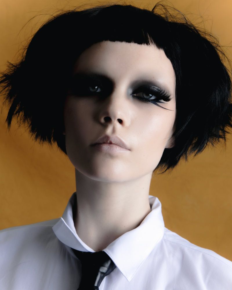Portrait of a model with short black hair and heavy dark smokey eye makeup wearing a white collared shirt and black tie.
