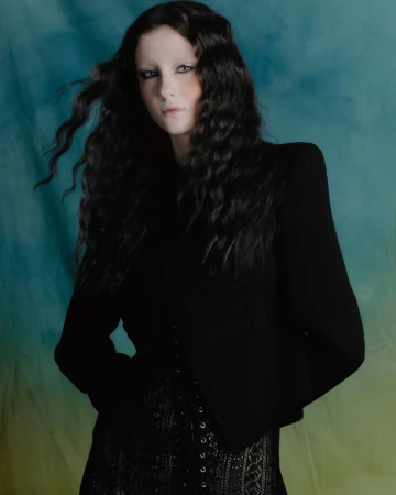 Portrait of a woman with long dark wavy hair wearing a black jacket and lace corset top against a blue-green backdrop.