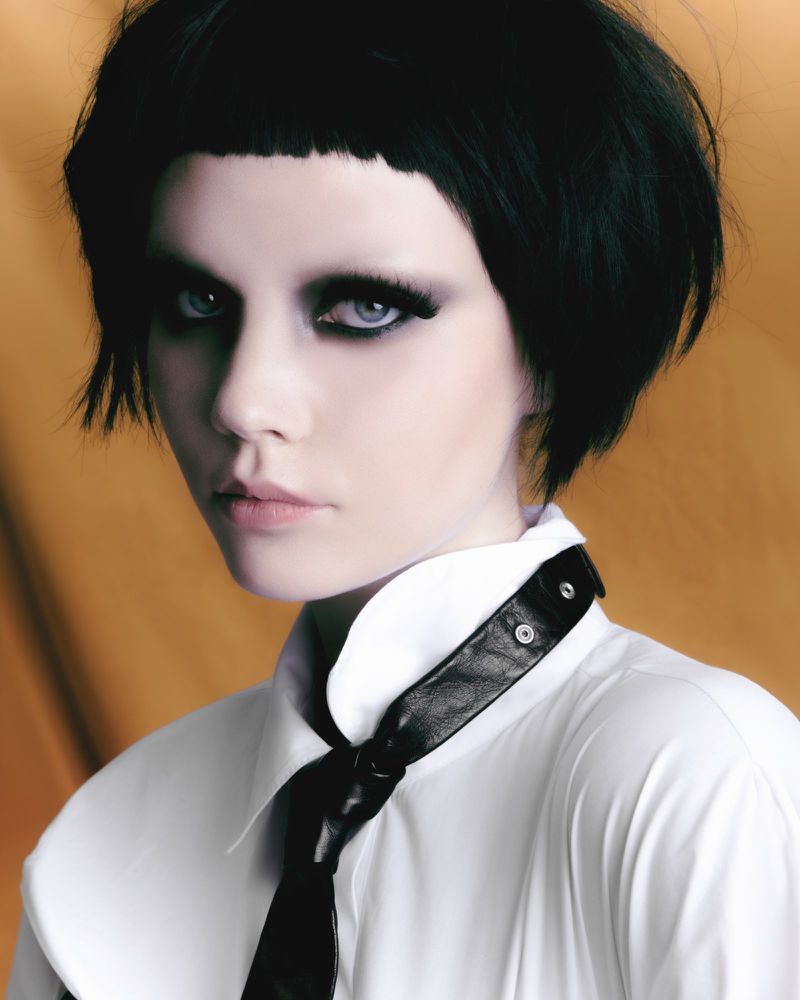 Close-up portrait of a woman with dark short hair and heavy smokey eye makeup wearing a white shirt and black necktie.
