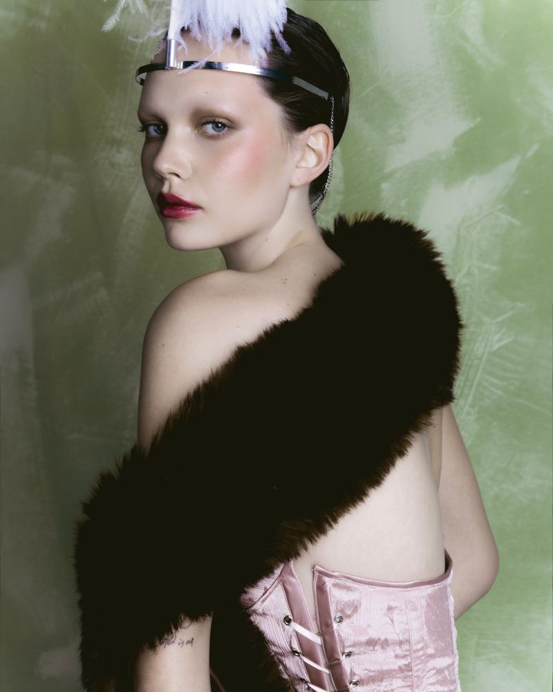 Model wearing a pink corset, dark fur stole, and feathered headpiece looking over shoulder against a green textured backdrop.
