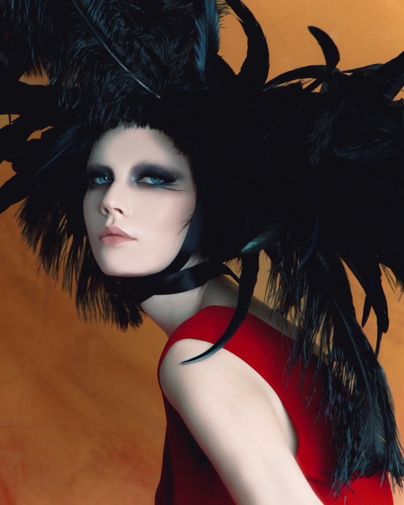 Model with dramatic smoky eye makeup wearing a large black feather headpiece and a red sleeveless garment against an orange background.