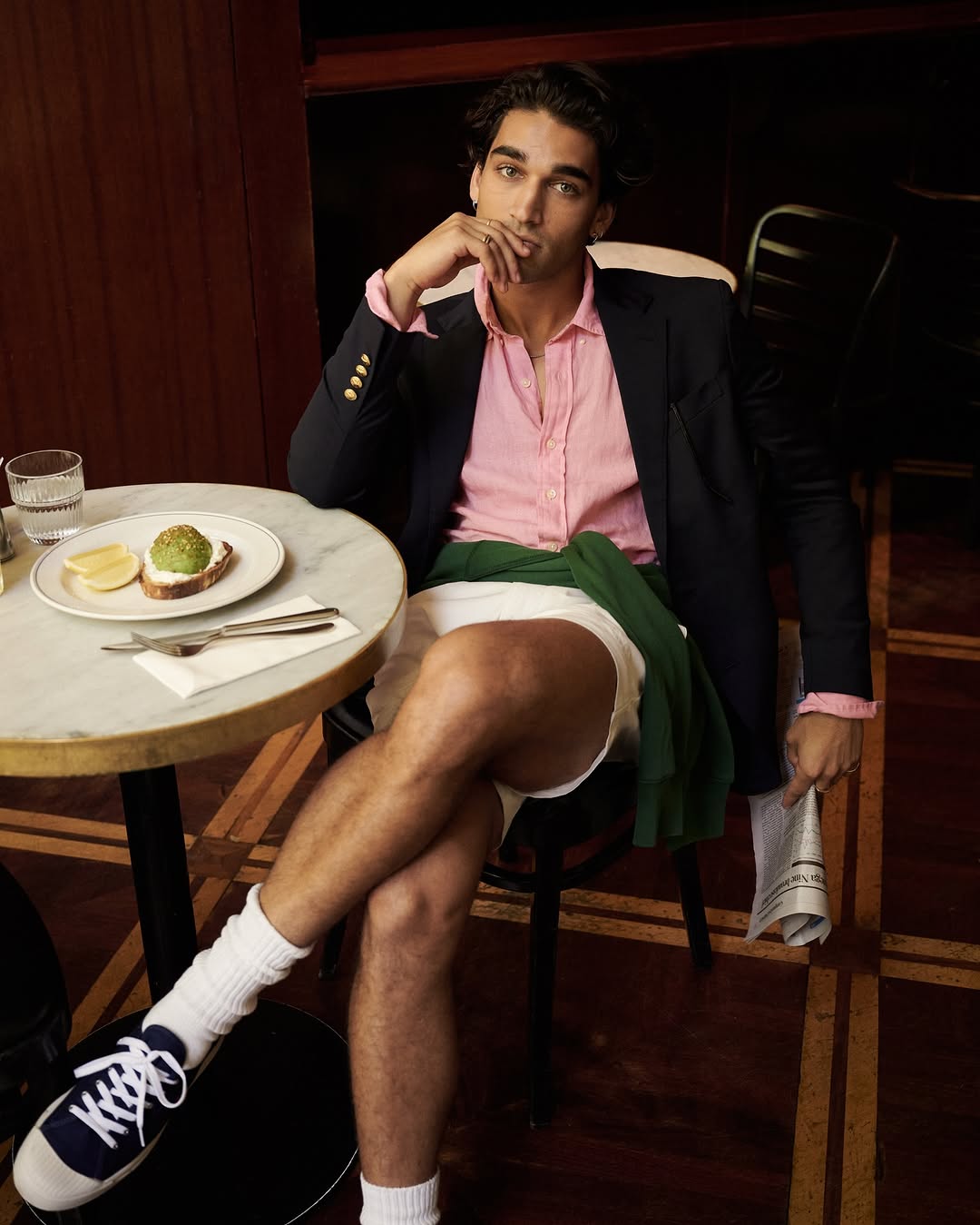 Man in preppy fashion sits at cafe with avocado toast, wearing blazer, pink shirt, shorts, socks, and sneakers in relaxed pose.