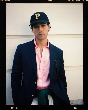 Man in navy blazer and cap with the letter P poses in front of an off-white wall. He's wearing a pink shirt and green sweater.
