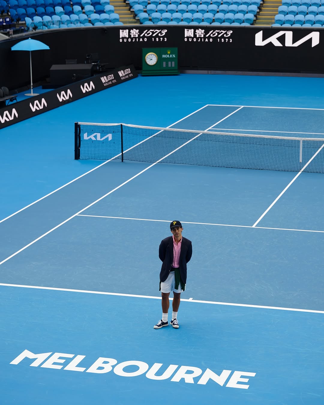 A man in a blazer stands on a blue tennis court at Melbourne Park, with logos, signage, and stadium seating in the background on a sunny day.