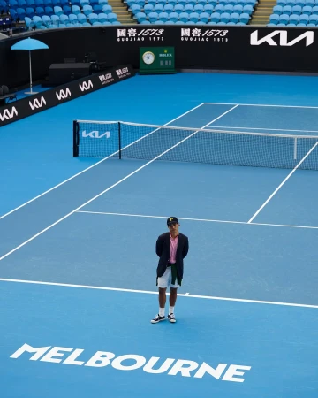 A man in a blazer stands on a blue tennis court at Melbourne Park, with logos, signage, and stadium seating in the background on a sunny day.