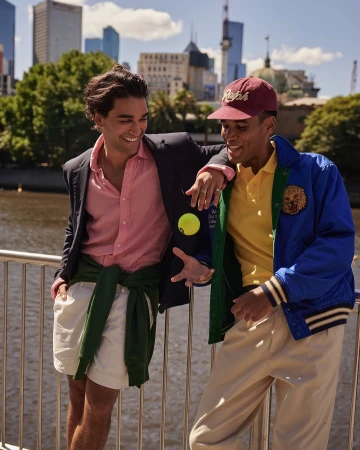 Two stylish men in casual wear stand near a waterfront in an urban setting, one holding a tennis ball, both smiling and looking at each other.