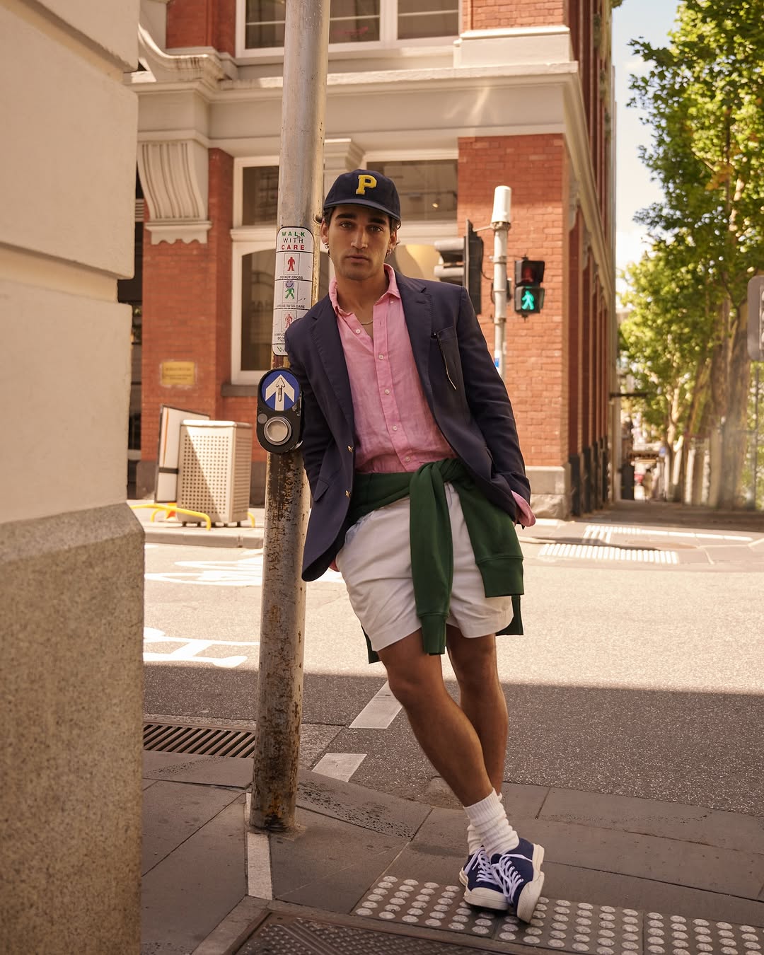 A young adult male leans against a traffic signal post in an urban street setting, wearing a blazer, shirt, shorts, cap, and sneakers.