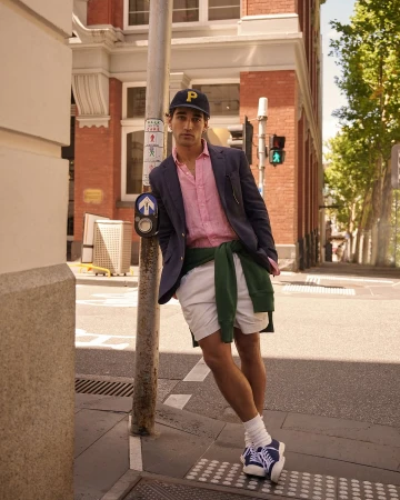 A young adult male leans against a traffic signal post in an urban street setting, wearing a blazer, shirt, shorts, cap, and sneakers.