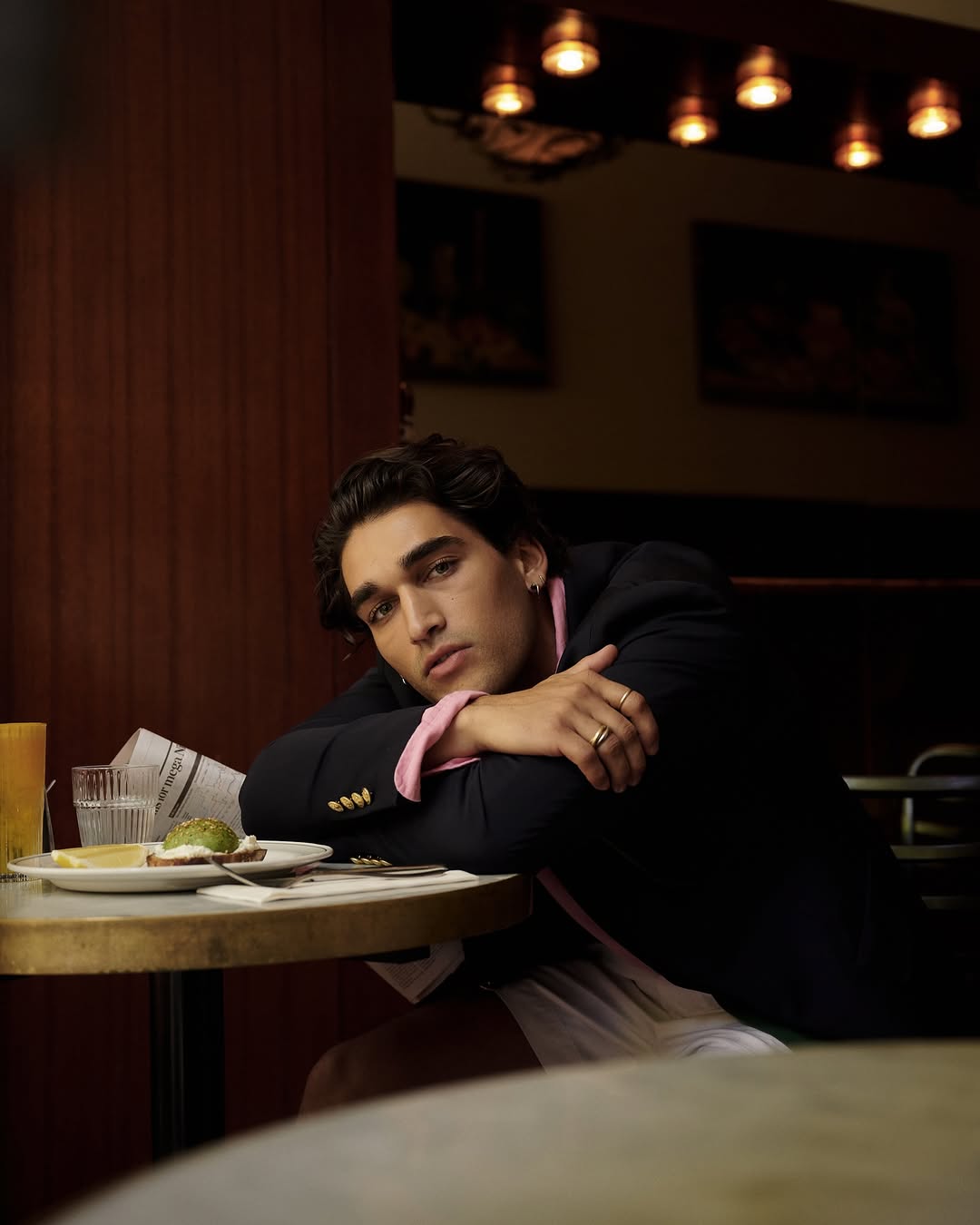 Adult man in a blazer sits at a table in a cafe with a drink, plate, and newspaper in the frame, softly lit indoors.