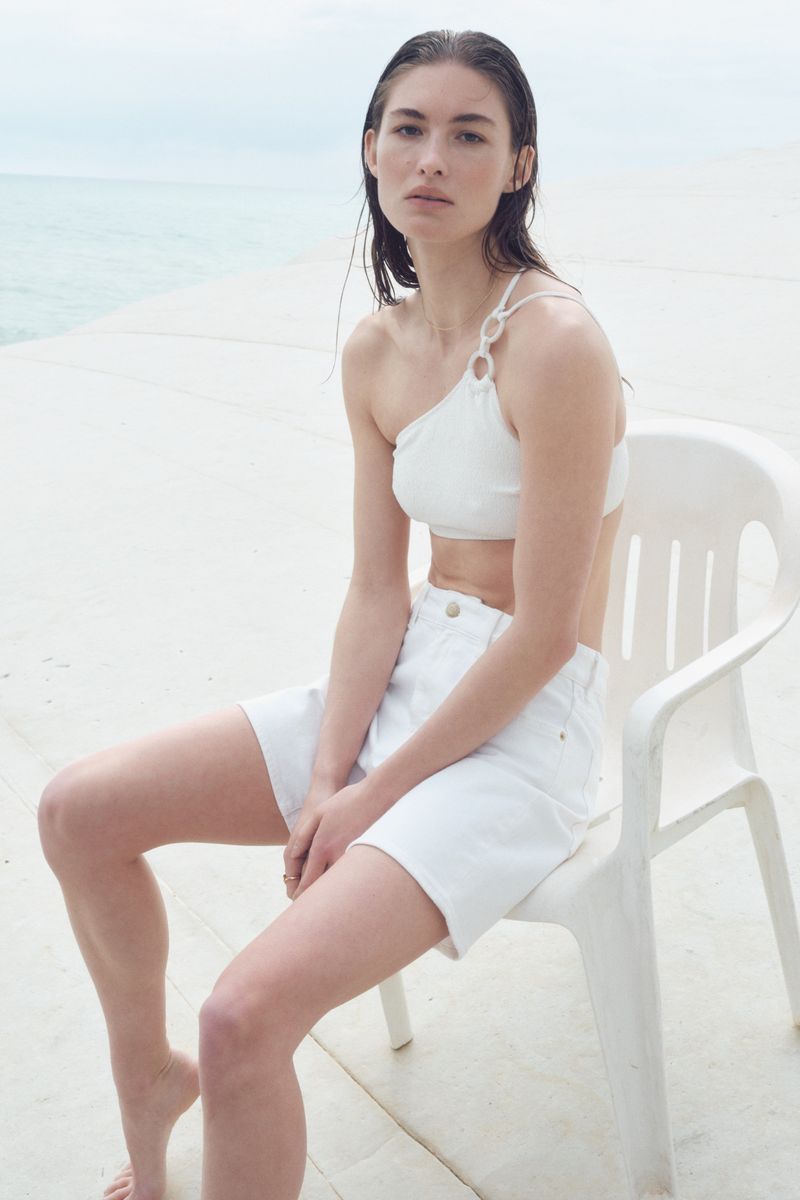 A woman with wet hair sitting in an outdoor setting, wearing a white bikini top and denim shorts against a light blue sky and ocean backdrop.