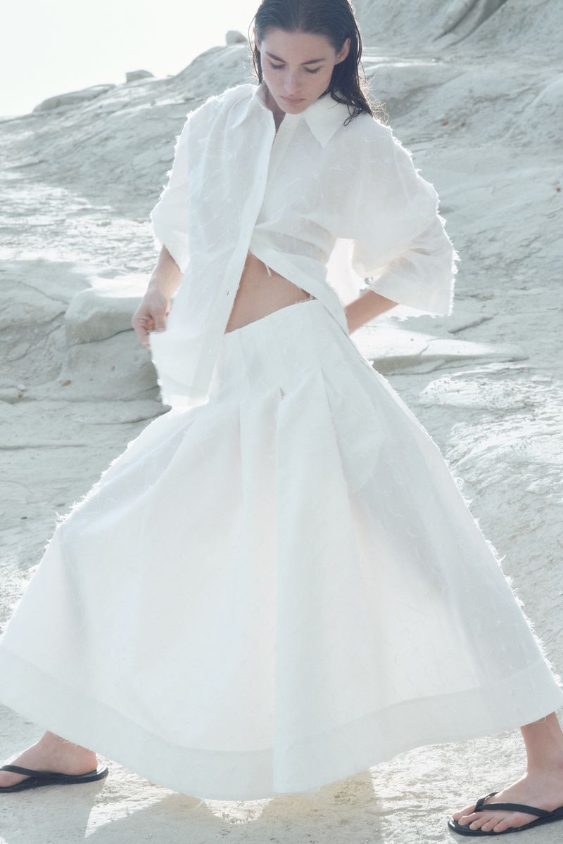 Fashion shot of a woman in a white linen shirt and matching long skirt, standing on a rock formation in bright outdoor lighting.