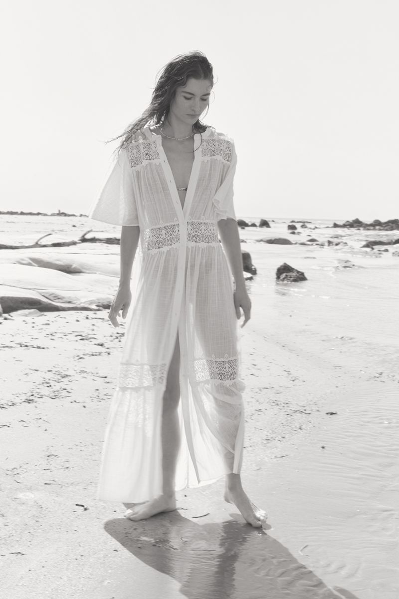 Woman in sheer white dress walks on a sandy beach with scattered rocks, enjoying a bright, sunny day near the water's edge.