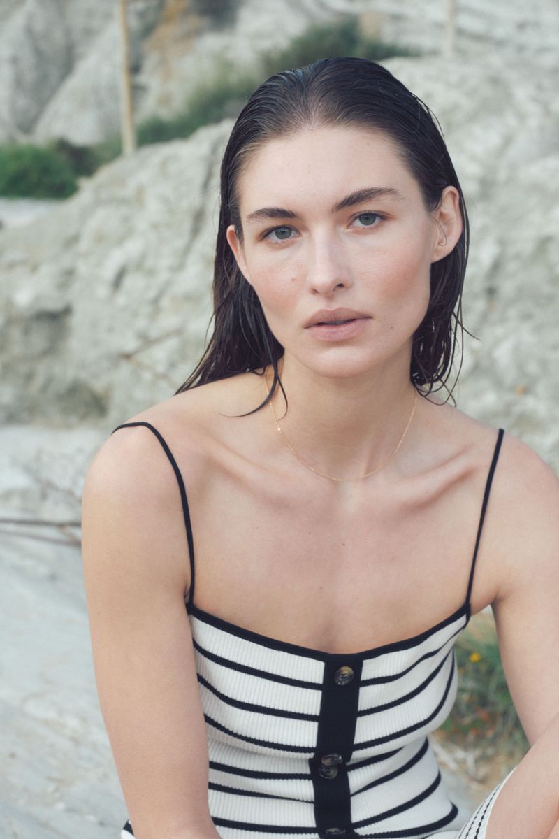 Portrait of a female model with wet hair wearing a black and white striped top with a button-up front, posing outdoors.