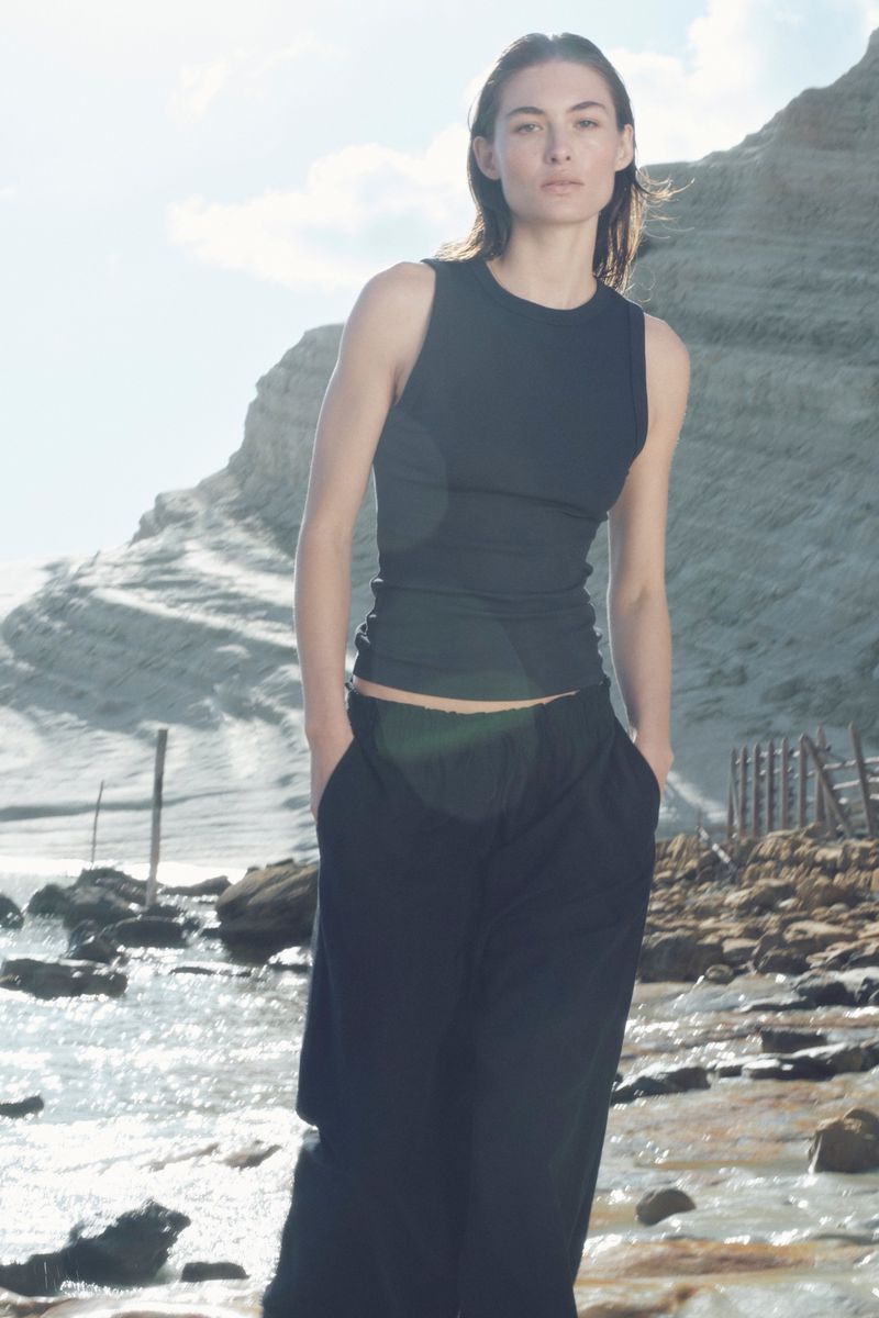 A young woman in a black tank top and pants stands on a rocky beach with a bright sky and pale cliffs in the background on a sunny day.