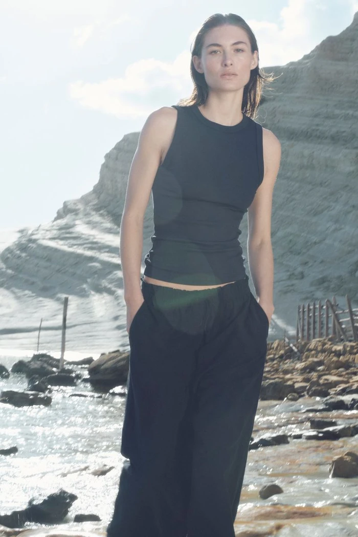 Grace Elizabeth standing on a rocky coast wearing a black sleeveless top and wide-leg trousers for H&M.