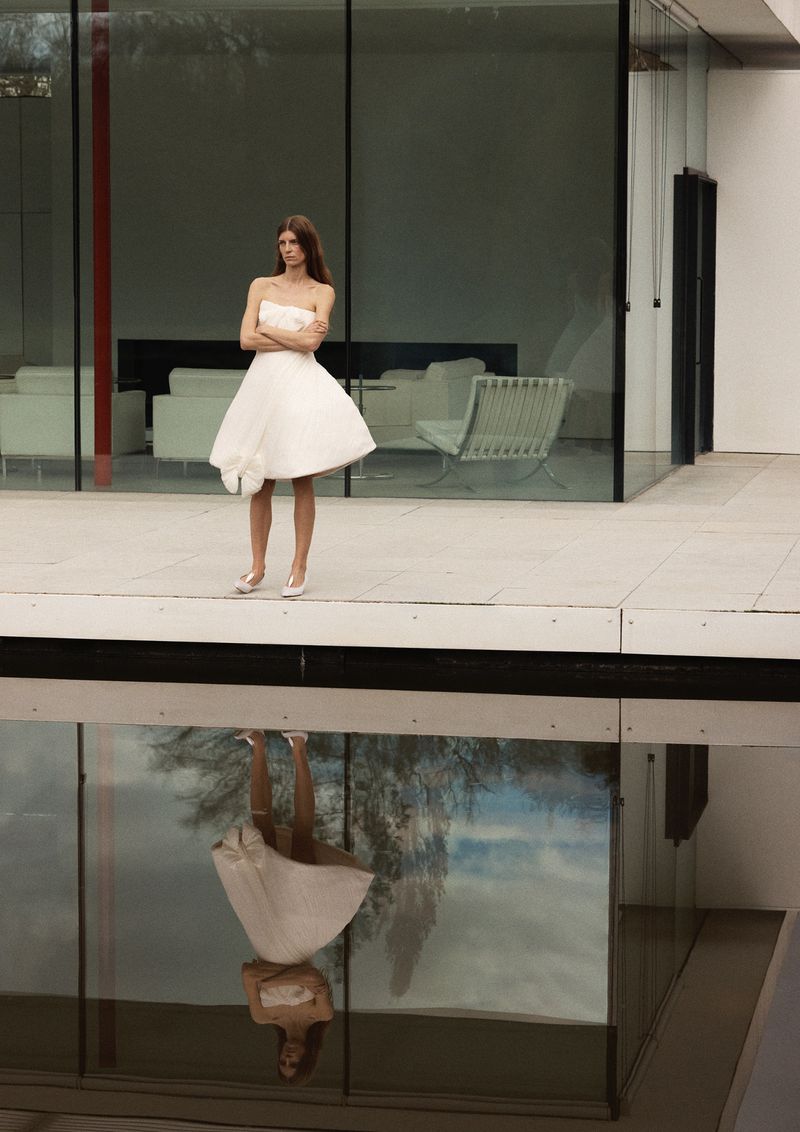 Model Luca Gajdus in a white strapless flared mini dress standing above a reflecting pool in front of a modern glass pavilion.
