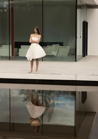 Model Luca Gajdus in a white strapless flared mini dress standing above a reflecting pool in front of a modern glass pavilion.
