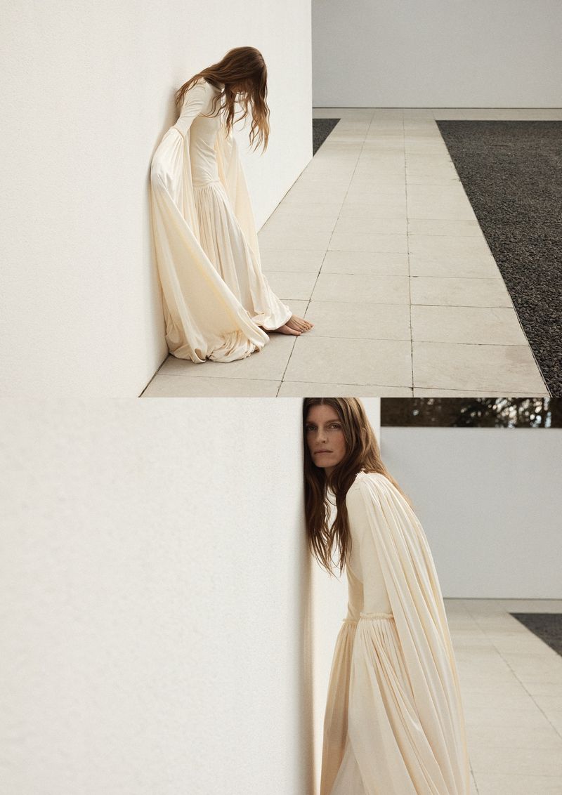 Model Luca Gajdus leaning against a minimalist white wall wearing a flowing cream draped gown in an outdoor courtyard setting.