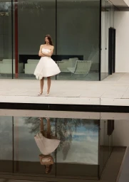 Model Luca Gajdus in a white strapless flared mini dress standing above a reflecting pool in front of a modern glass pavilion.