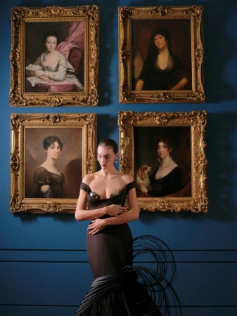Model in a black off-the-shoulder gown standing in front of four framed classical portrait paintings on a blue wall.