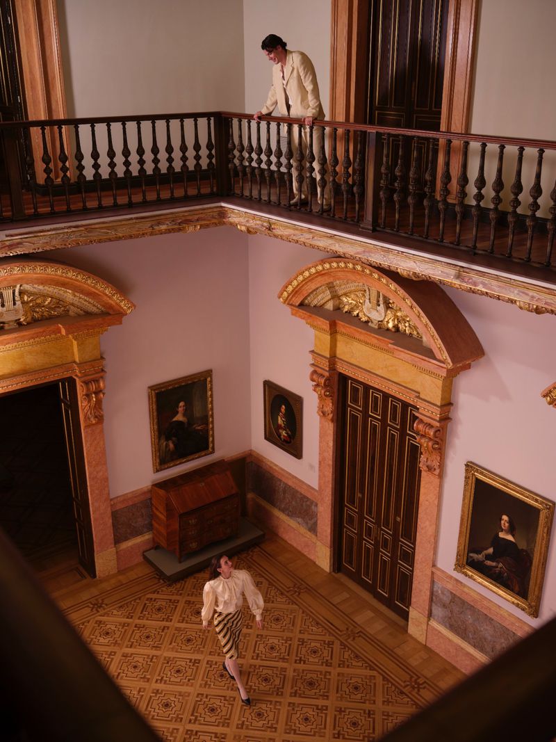 Two figures in a grand historic interior with pink walls, dark wood trim, and framed portraits on the walls.