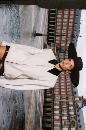 Model wearing an oversized wide-brim black hat and white collared zip jacket in an open historic European plaza.